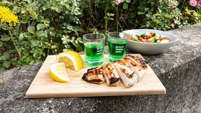 Das Bild zeigt eine gegrillte Hühnerbrust die aufgeschnitten auf einem Brettchen liegt neben einer Schüssel Salat, ein paar Pfeffi Pinnchen und einer Zitrone